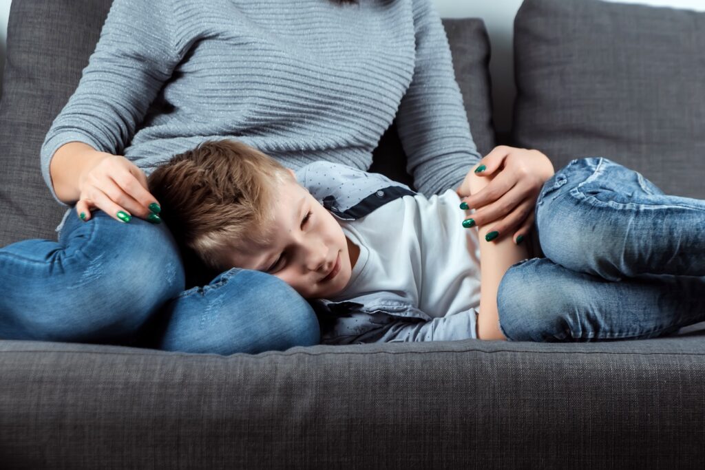 El niño está tumbado en el sofá con dolor abdominal cerca de su madre. El concepto de custodia, cuidado parental, problemas estomacales, intoxicación alimentaria, problemas infantiles.